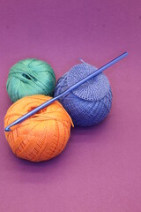 balls of knitting threads green orange and blue lie on a purple background and a blue crochet hook on top