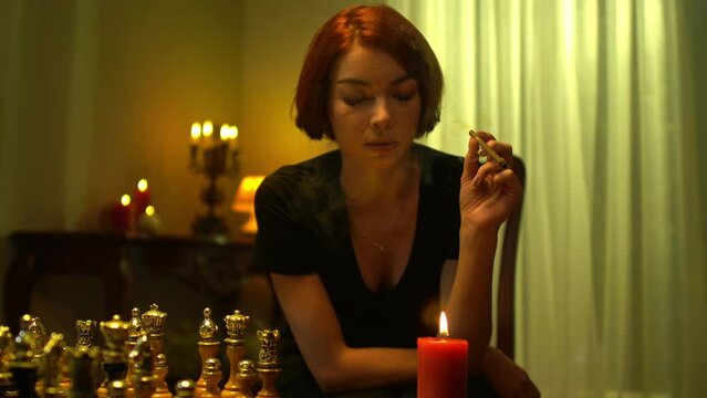 Redhead beautiful woman lighting cigar from candle sitting at table with chess board indoors. Portrait of Caucasian chess player thinking smoking planning game strategy