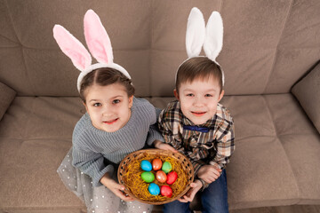 A girl and a boy in bunny ears hold a wicker basket with Easter colored eggs and look up at the camera while sitting on the couch. Children