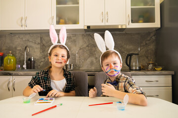 A girl and a boy in a headband with bunny ears are sitting at the kitchen table with painted faces and looking at the camera