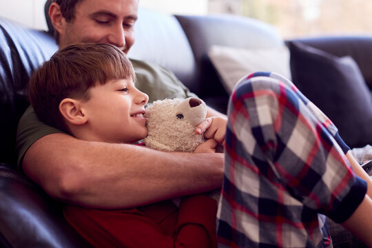 Father Cuddling Son With Toy Dog Sitting On Sofa In Pyjamas Together