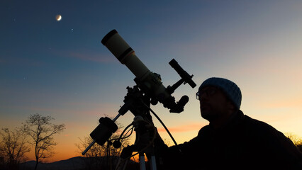 Silhouette of a man, telescope and countryside under evening skies.