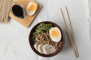 Asian food ramen noodles with meat, vegetables and egg in a brown bowl on a light background, soy sauce in a gravy boat, chopsticks