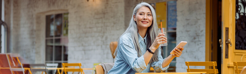 Elegant grey haired Asian woman with glass of water and smartphone sits at small table on outdoors...
