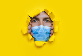 A man wearing a protective medical mask that completely covers his face peeps through a torn hole in yellow paper. The concept of a pandemic,quarantine and protection against coronavirus and covid-19.