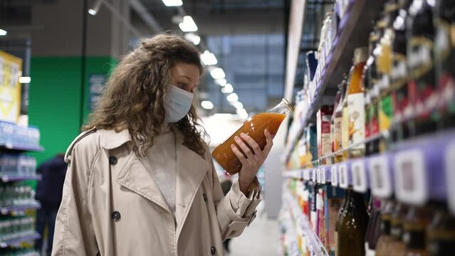 Woman in trench coat chooses fresh juice reading ingredients on glass bottle in supermarket. Young customer with medical mask at shopping during pandemics