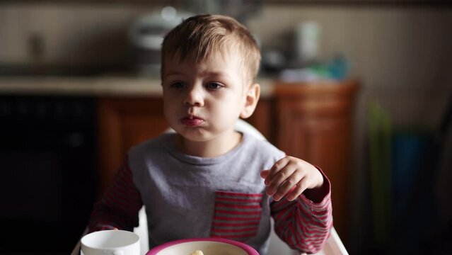 A 3-year-old boy coughed and eats biscuits while sitting behind a chair.