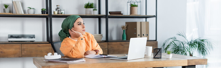 muslim woman looking away while sitting near laptop at home, banner.