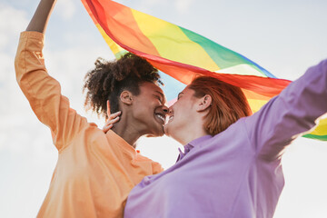 Mulitracial lesbian girls kissing together at gay pride parade while holding rainbow flag - LGBT concept - obrazy, fototapety, plakaty