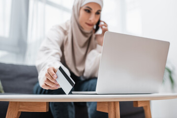 Fototapeta premium blurred arabian woman talking on mobile phone while holding credit card near laptop.