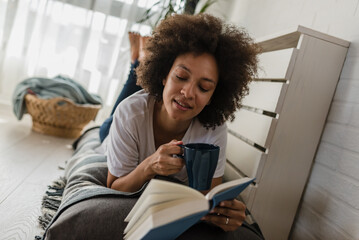 Beautiful African-American young Woman reading a book at home. Cozy corner. Education. Daydreaming and passion concept
