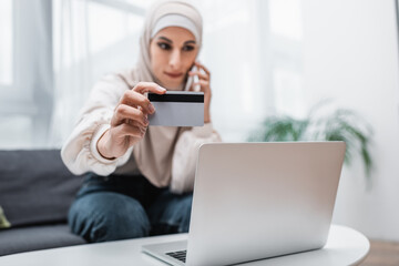 selective focus of credit card in hand of blurred arabian woman calling on smartphone near laptop.