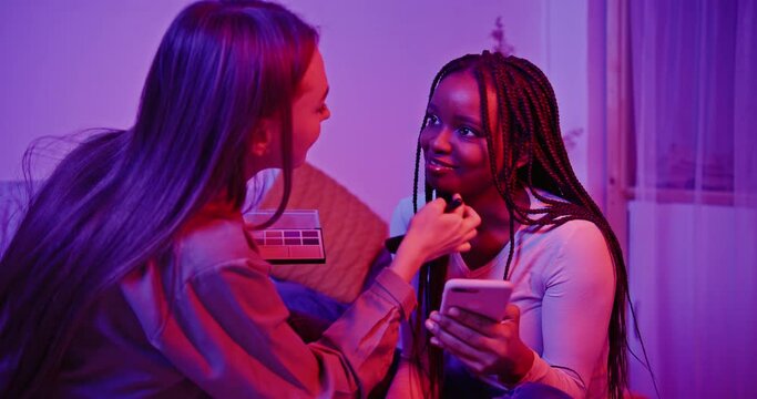 Diverse girls doing makeup and browsing smartphone. Cheerful multiracial teenage best friends applying makeup while surfing social media on mobile phone and having fun in dark room with pink neon