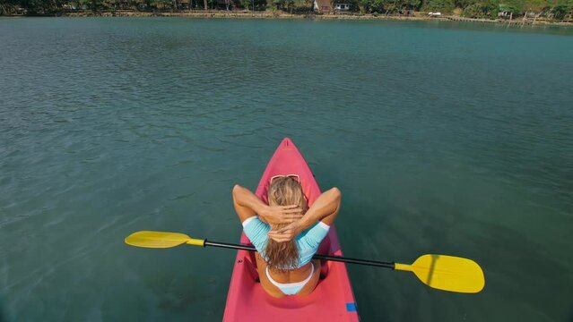 Seductive blonde woman in blue swimsuit sails on pink plastic kayak along azure sea at exotic resort backside upper above view. Traveling to tropical countries. Girl is sailing on kayak in ocean.