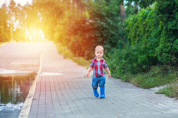 Toddler boy in rubber boots lies and runs through puddles after rain..