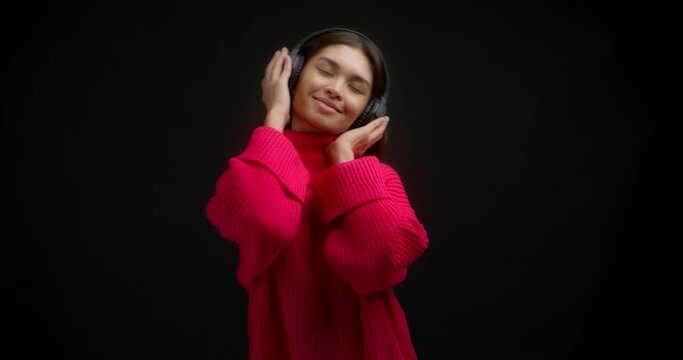 Beautiful asian woman in a bright pink sweater enjoys music in wireless headphones. Cute woman dancing to the song isolated on a black background.
