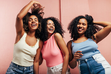 Three young women in bright casuals dancing together at a pink wall and having fun
