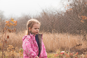 Little beautiful cute sad girl in a jacket with blond hair in pigtails stands on an autumn background