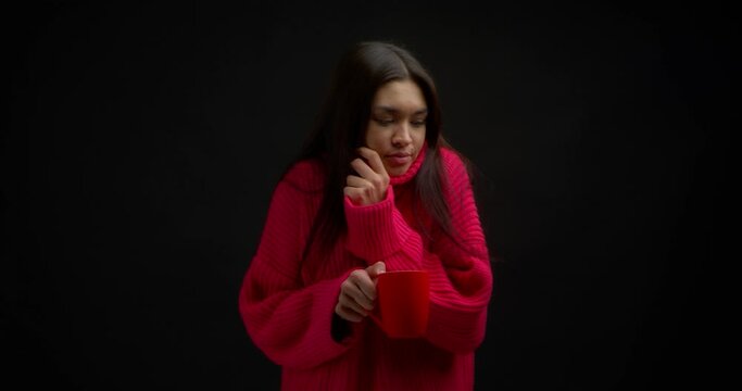 Beautiful brunette with long hair in a pink sweater froze. Asian woman has chills, warms up with a hot drink in a red mug. Isolated on a black background.