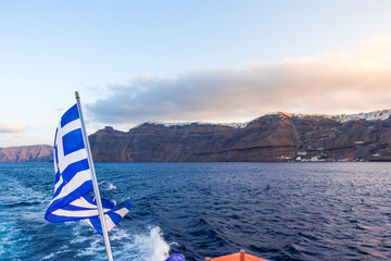 Cruise ship anchor in Aegean sea, Santorini island, Thera city view from speedboat with Greek flag. Greece.
