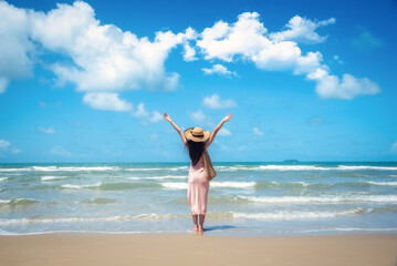 Happy traveler woman in sunhat and dress standing with her arms raised looking tropical beach on summer day. Holiday and summer travel concept