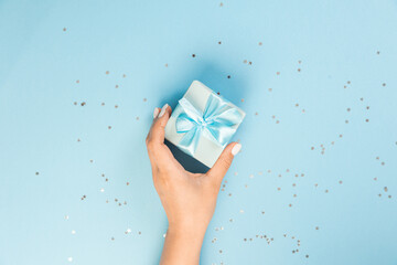 A hand holds a blue gift box on a blue background with glitter stars. top view, copy space