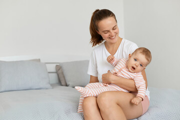 Horizontal shot of young adult happy mother wearing white casual t shirt sitting on bed in bedroom with toddler baby, mommy looking at child with charming smile, being glad to spend time with her kid.