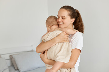 Indoor shot of happy satisfied woman holding daughter wrapped in towel after bathroom, lovely mother hugging her toddler baby with love, looking after kid.