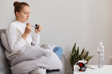 Indoor shot of sick female wearing white sweater and bun hairstyle, sitting on sofa wrapped in...