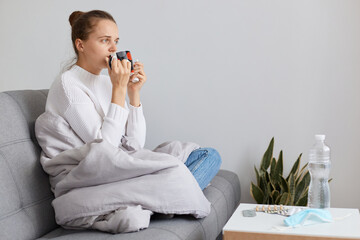 Indoor shot of unhealthy female wearing white sweater and bun hairstyle, sitting on sofa wrapped in blanket and drinking hot tea, having headache and high temperature.