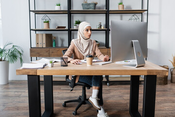 full length view of arabian woman working on computer near takeaway drink and documents on desk.