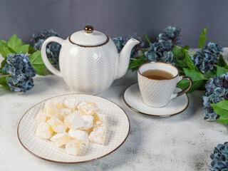 A teapot and a white mug on the table, flowers and traditional Turkish and Greek jelly candies in powdered sugar on a plate. confectionery in Arabic style..T The concept of recipes, 