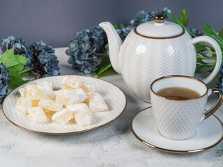A teapot and a white mug on the table, flowers and traditional Turkish and Greek jelly candies in powdered sugar on a plate. confectionery in Arabic style..T The concept of recipes, 