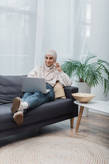 cheerful arabian woman in hijab eating popcorn and watching film on laptop on couch.