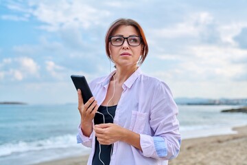 Middle age sad serious woman in headphones with smartphone outdoor.