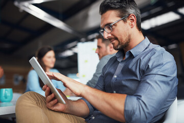 Letting technology do the work for him. Shot of a man sitting at a table in an office using a digital tablet with colleagues working in the background.