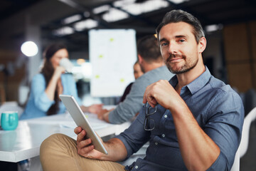 Bringing digital ideas to the meeting. Portrait of a man sitting at a table in an office using a digital tablet with colleagues working in the background.