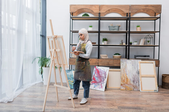 full length view of muslim woman drawing on easel near painted pictures in home studio.