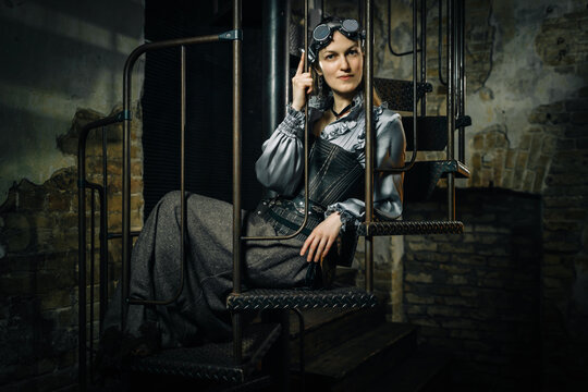 A girl in a steampunk costume sits on a metal ladder against a brick wall