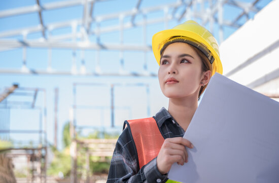 Asian woman civil engineer wear yellow safety hard hat standing and holding blueprint while checking infrastructure of building in construction site.