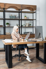 muslim woman looking at document while sitting near computer at home.
