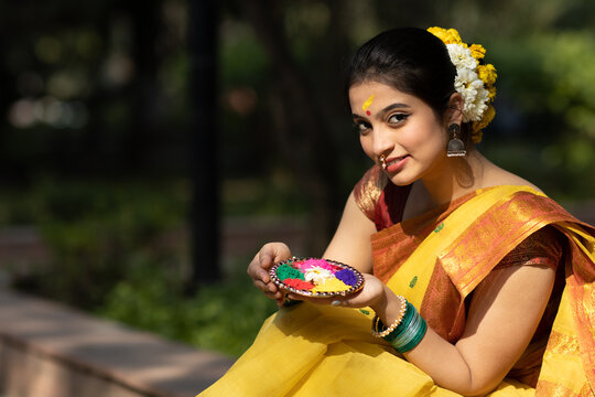 Beautiful and young girl or woman in yellow saree seating in garden with a decorated plate of color or colour or gulal or abeer or Holi powder to celebrate holi festival of colors