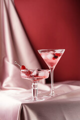 Martini and champagne glasses with berry cocktail drink with ice cubes and crashed ice on pink and burgundy background