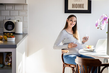 Pregnant woman sitting at table at home eating fruit salad and using laptop.