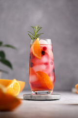 A tall glass with cold orange juice with grapefruit, frozen berries and ice cubes, rosemary, grapefruit half on grey background