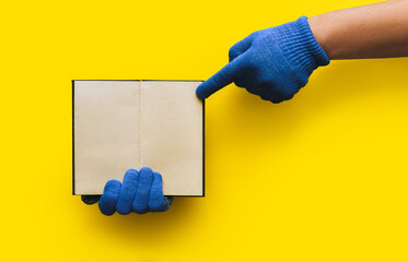 The hands of a worker in blue knitted gloves hold a notebook on a yellow background and points to...