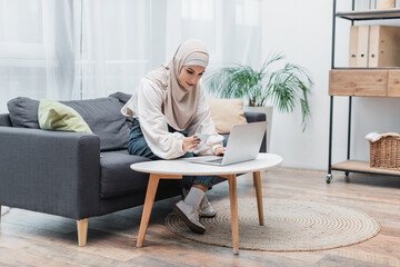 Fototapeta premium muslim woman in hijab holding credit card and using laptop on couch at home.