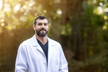 A doctor with dark hair and a beard in a white lab coat standing outside in a natural green environment