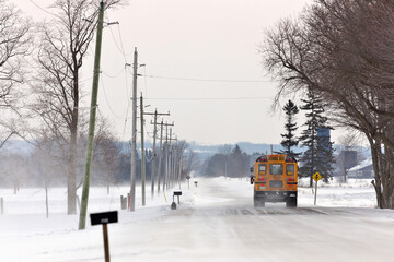 Fototapeta premium School Bus Travelling Down a Country Road with Snowdrifts and Blowing Snow in Winter