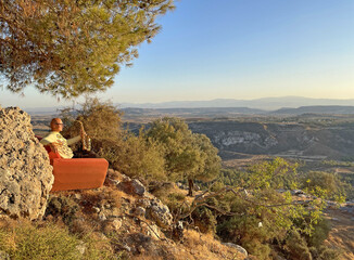 Young stylish bald male saxophonist sits on an orange sofa in mountains of Cyprus in nature, under pine tree, holds an alto saxophone in his hands. Musician with saxophone on nature in mountains